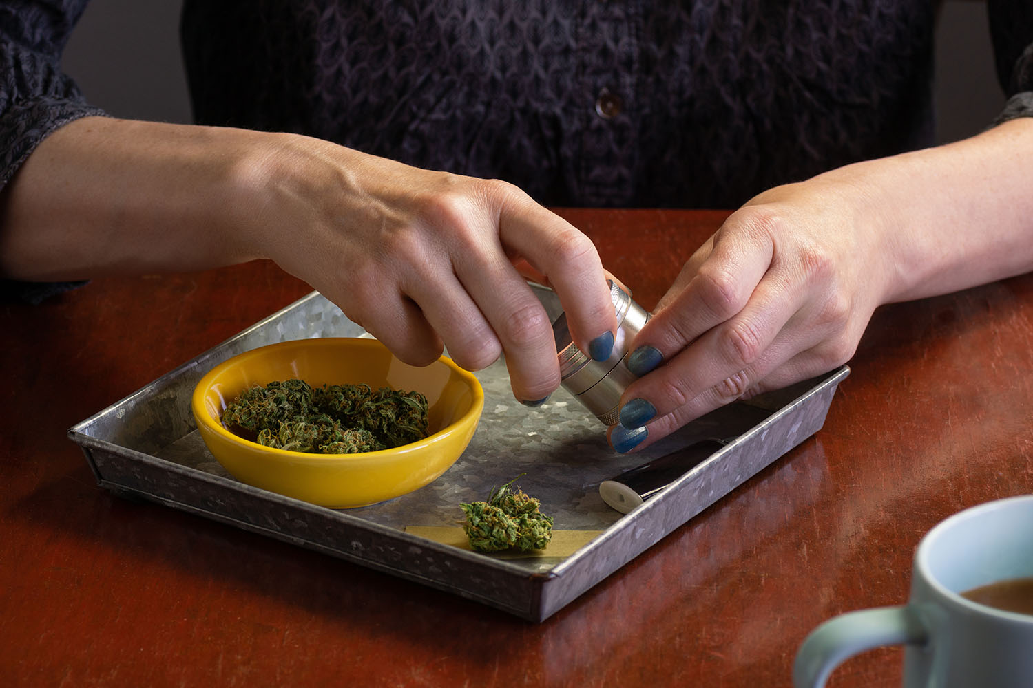 Hands turning a marijuana grinder over a metal rolling tray holding a yellow bowl of marijuana flower